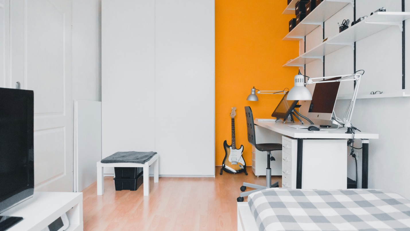 A bedroom with an orange accent wall, white shelving, and a desk with a computer. 